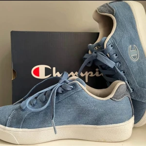 Blå sneakers i canvas från Champion - Snygga blå sneakers från Champion i canvasmaterial med vita detaljer och snörning. Skorna har rund tå, platt sula och diskret logga på sidan. Perfekta för dig som gillar en clean och avslappnad stil.