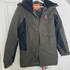 Fjällräven jacka  - Säljer en grön Fjällräven jacka i storlek XS. Jackan har svarta detaljer på sidorna och är utrustad med flera stora fickor framtill, huva och dragkedja. 