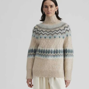 Beige mönstrad polotröja By Malina  - Supermysig varm beige polotröja från By Malina med stickat mönster i blått, grått och vitt. Tröjan har hög krage, lång ärm och ribbade muddar. Materialmix med ull, mohair och återvunnen polyester ger en fluffig och varm känsla. Perfekt för kalla dagar! Inga anmärkningar, mycket fint skick utan noppor.