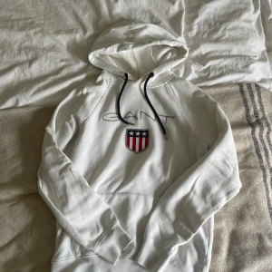 Vit hoodie från Gant  - Vit hoodie från Gant med broderad logga och amerikanskt inspirerat emblem på bröstet. Tröjan har huva med svarta snören och klassisk känguruficka. Perfekt för en avslappnad och stilren look.
