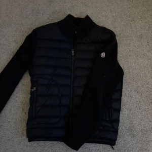 Svart Moncler pufferjacka med dragkedja - Svart pufferjacka från Moncler med quiltad front, ribbad krage och ärmar. Jackan har två fickor med dragkedja och klassisk Moncler-logga på ärmen. Perfekt för kyliga dagar och har en stilren, sportig look.