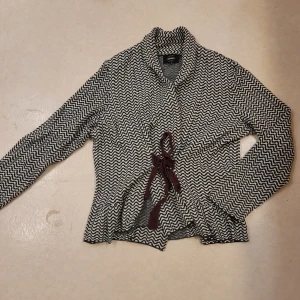 Svartvit kofta från Only med knyt - Säljer en svartvit mönstrad kofta från Only med vågigt mönster och snyggt knytband i midjan. Koftan har lång ärm och en skön, mjuk känsla. Perfekt för lager-på-lager och att slänga över en topp.