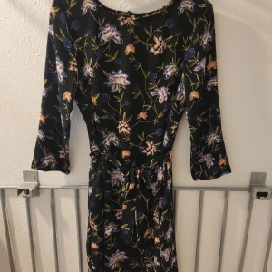 Svart blommig klänning med knytband - Svart klänning med färgglatt blommönster och trekvartsärmar. Klänningen har ett knytband i midjan som ger en snygg siluett. Perfekt för dig som gillar en feminin och trendig stil.