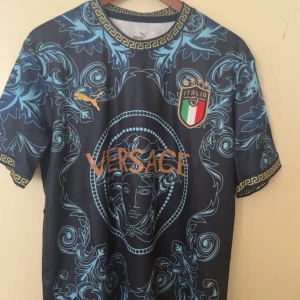 Blå/guld fotbollströja - Unik fotbollströja med klassiskt Medusa-motiv. Mörkblå bas med ljusblå barockmönster, gulddetaljer och grekisk bård runt hals och ärmslut. Tillverkad i lätt, glansigt material. Perfekt för dig som vill sticka ut på planen.