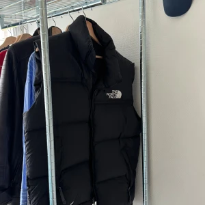 Svart dunväst från The North Face - Säljer en svart dunväst från The North Face med hög krage och dragkedja framtill. Västen har en klassisk puffer-look och logga broderad på bröstet. Perfekt för lager-på-lager och streetstyle.