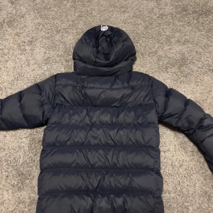 Mörkblå dunjacka Sail Racing - Säljer en mörkblå dunjacka från Sail Racing med huva och dragkedja framtill. Jackan är quiltad och har en clean look med logga på bröstet. Perfekt för kalla dagar när du vill hålla värmen och ändå se snygg ut.