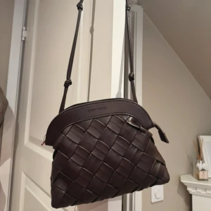 Quiltad väska - Snygg brun axelväska med flätad design och justerbar axelrem. Väskan är tillverkad i skinnimitation och har en dragkedja upptill. Perfekt storlek för det viktigaste och ger en trendig touch till din outfit.