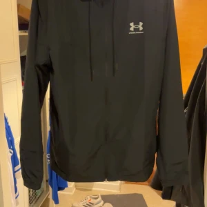 Svart vindjacka från Under Armour M - Svart vindjacka från Under Armour med huva och dragkedja framtill. Jackan har en clean look med logga på bröstet och justerbar dragsko i huvan. Perfekt för blåsiga dagar och sportiga outfits.
