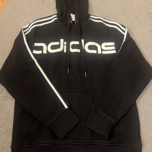 Svart Adidas hoodie med vita ränder - Svart hoodie från Adidas med klassiska vita ränder längs axlarna och stort vitt logotryck över bröstet. Tröjan har huva med snörning och en stor magficka. Perfekt för en sportig och avslappnad stil.