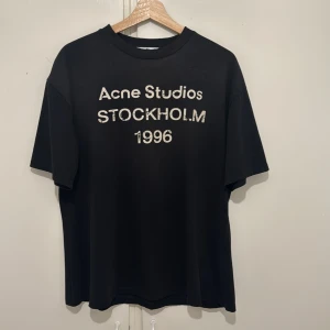 Svart Acne Studios t-shirt Stockholm - Svart t-shirt från Acne Studios med tryck 'Acne Studios STOCKHOLM 1996' på bröstet. Klassisk passform med rund hals och korta ärmar. Tillverkad i 100% ekologisk bomull för en skön och mjuk känsla. Perfekt för en clean och stilren look.