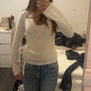 Vit omlottkofta med knytband - Mysig vit omlottkofta med knytband i sidan och v-ringad hals. Koftan har långa ärmar och är stickad i ett mjukt material. Perfekt att slänga på sig över ett linne eller t-shirt för en chill och stilren look.