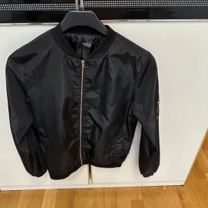 Svart bomberjacka  - Svart bomberjacka  med klassisk ribbad krage, muddar och dragkedja framtill. Jackan har en stilren look med ficka på ärmen och är tillverkad i ett glansigt syntetmaterial. Perfekt för dig som vill ha en clean och tidlös stil.