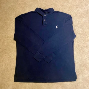 Marinblå långärmad pikétröja Polo Ralph Lauren - Säljer en marinblå långärmad pikétröja från Polo Ralph Lauren med vit broderad logga på bröstet. Tröjan har klassisk krage, knäppning med två knappar och är i bomull. Slim fit-modell som sitter snyggt och stilrent.