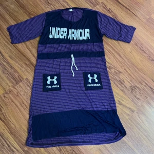 Randig lila klänning från Under Armour - Sportig lila och marinblå klänning från Under Armour med svarta och vita ränder, knytband i midjan och två stora fickor framtill med logga. Halvlånga ärmar och avslappnad passform, perfekt för en aktiv stil.