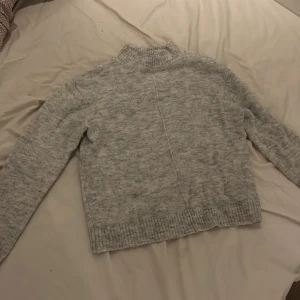 Grå stickad tröja från K by Kappahl - Mysig grå stickad tröja från K by Kappahl i storlek XS. Tröjan har en hög, ribbad krage och långa ärmar. Perfekt för kyliga dagar och enkel att matcha med jeans eller kjol.