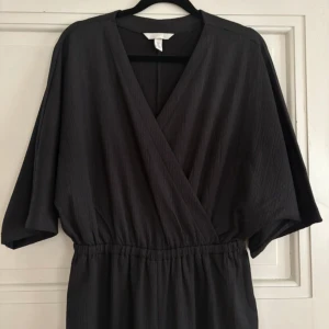 Svart jumpsuit från H&M - Svart jumpsuit från H&M i mjuk polyester med omlott framtill, resår i midjan och vida, korta ärmar. Jumpsuiten har en stilren look. Perfekt för dig som gillar minimalistisk och avslappnad stil.