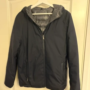 Mörkblå vändbar Ralph Lauren jacka med huva - Snygg vändbar pufferjacka från Polo Ralph Lauren i mörkblått och silvergrått. Jackan har huva, dragkedja framtill och två sidofickor med dragkedja. Ena sidan är quiltad och glansig, andra sidan är matt och stilren. Perfekt för kalla dagar.