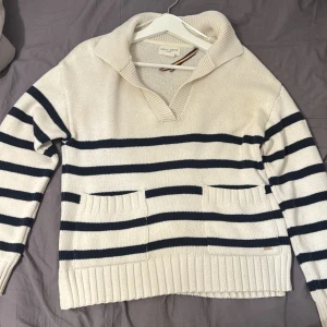 Randig stickad tröja från Holly & White - Säljer en krämvit stickad tröja från Holebrook med marinblå ränder, djup v-ringning och bred krage. Tröjan har två fickor framtill och ribbade muddar. Perfekt för en chill och stilren look.