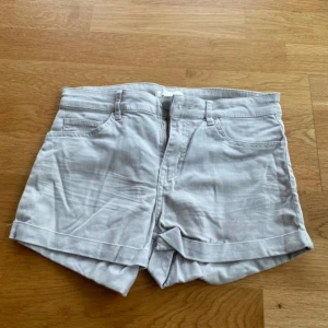 Beige denimshorts med uppvikt kant - Beige shorts i denim med klassisk femficksdesign och uppvikta benslut. De har bälteshällor, knapp och dragkedja framtill samt bakfickor. 
