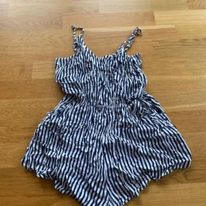 Randig kort playsuit - Kort platsuit med smala axelband och blåvita ränder. Klänningen har resår i midjan. 