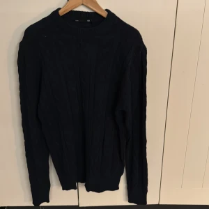 Mörkblå stickad tröja från 157 - Mörkblå stickad tröja från 157 med klassiskt kabelstickat mönster. Tröjan har rund halsringning och långa ärmar, perfekt för lager-på-lager under kyliga dagar. Enkel och stilren design som funkar till det mesta.