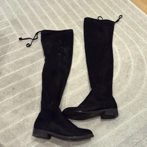 Svarta overknee stövlar i mocka - Säljer ett par svarta overknee stövlar i mjuk mocka med rund tå och platt sula. Stövlarna har dragkedja på insidan och knytning upptill för extra passform. 
