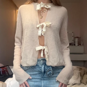 Beigerosa kofta med rosettknäppning - Superfin beigerosa kofta med tre stora rosetter framtill som knäppning. Långärmad och i mjukt stickat material, perfekt för lager-på-lager. Snygg och trendig detalj med de breda knytbanden framtill. Passar till både jeans och kjol.