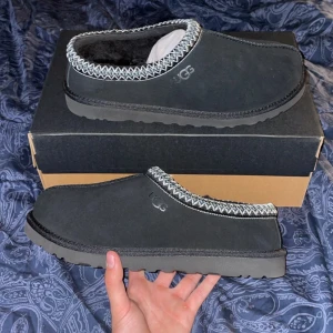 Svarta UGG slip-on boots - Svarta slip-on boots från UGG TASMAN med mjuk insida och grov sula. Skorna har ett snyggt mönster i vitt och grått runt öppningen och är tillverkade i mocka med fårullsfoder. Perfekta för kalla dagar och riktigt sköna att glida i. Helt nya oanvända!
