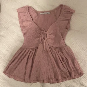 Rosa blus med volang och knyt - Supersöt ljusrosa blus från  lager 157 i storlek XS. Blusen har volangdetaljer vid axlarna, v-ringad hals och knyt framtill. Tillverkad i ett mjukt, krinklat material. 