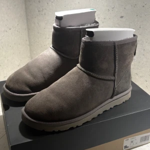 Grå UGG boots i mocka - Säljer ett par klassiska UGG boots i grå mocka med rund tå och platt sula. Skorna har en mjuk insida och tydlig UGG-logga baktill. Perfekt för kalla dagar och har en robust yttersula med mönster för extra grepp. ”Använda max 10 gånger men finns små tecken på användning”