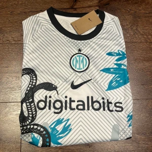 Inter Milan Nike fotbollströja Dri-FIT - Inter Milan fotbollströja från Nike med Dri-FIT-material. Vit bas med grått geometriskt mönster, svarta detaljer och turkosa inslag. Tryckt ormmotiv och klubbmärke på bröstet. Svart krage och ärmslut. Snygg och modern design för alla Inter-fans.