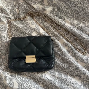 Svart axelväska från H&M - Snygg svart quiltad axelväska från H&M med guldfärgad kedja och lås. Väskan är i mjukt syntetmaterial och har ett klassiskt rutmönster. Perfekt storlek för det viktigaste och ger en trendig vibe till din outfit. 