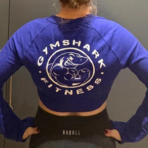 Blå croppad långärmad Gymshark-tröja - Säljer en blå croppad långärmad tröja från Gymshark med stort tryck på ryggen och tre knappar framtill. Perfekt för gymmet eller träning, i mjuk bomullskvalitet och med normal passform. Snygg och sportig vibe! Endast testad! 