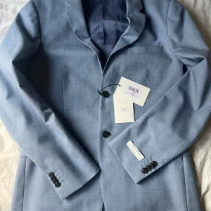 Ljusblå kavaj Tiger of Sweden - Snygg ljusblå kavaj från Tiger of Sweden, modell Jerretts. Klassisk passform med två knappar framtill, fyra knappar vid ärmslut och diskreta fickor. Tillverkad i ull med slät yta och helfodrad insida. Perfekt för dig som vill ha en stilren och modern look. Kavajen är sprillans ny. Säljes till ett väldigt bra pris. (Nypris, 3700kr) prislapp finns kvar.