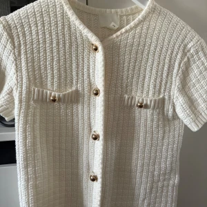Vit kortärmad stickad topp H&M - Superfin vit stickad topp från H&M med korta ärmar och snygga guldknappar framtill. Toppen har två dekorativa fickor på bröstet och en ribbad struktur som ger en lyxig känsla. Perfekt för dig som gillar klassisk och stilren look.