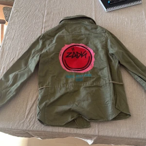 Olivgrön overshirt från Zadig & Voltaire - Snygg olivgrön overshirt från Zadig & Voltaire med coolt tryck på ryggen i rött och rosa samt texten 'Paris New-York 2022'. Jackan har klassisk krage, stora fickor framtill och är tillverkad i bomull. Perfekt för dig som vill sticka ut med streetstyle-vibe. En knapp som ramlat av på framsidan men annars i fint skick!