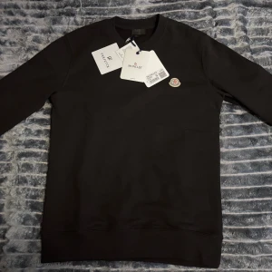 Moncler sweatshirt - Svart sweatshirt från Moncler i 100% bomull med klassisk rund hals och broderad logga på bröstet. Enkel och stilren design, perfekt för dig som gillar exklusiva basics. Tröjan har långa ärmar och ribbade muddar. Snygg att matcha med jeans eller joggers.