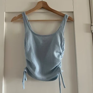 Ljusblå ribbad linnetopp med knyt S  - Supersöt ljusblå linnetopp med ribbad struktur och knyt i sidorna. Toppen är ärmlös med breda axelband och har en lätt croppad passform. Perfekt för varma dagar och enkel att styla till jeans eller kjol.