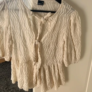Beige blus med knyt från Gina Tricot - Supersöt beige blus från Gina Tricot i storlek XS. Blusen har ett strukturerat tyg med vågigt mönster, korta puffärmar och två knyt framtill. Den har en luftig och flowy passform, perfekt för en chill och trendig look.