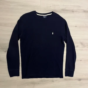 Mörkblå långärmad tröja Polo Ralph Lauren - Mörkblå långärmad tröja från Polo Ralph Lauren i 100% bomull. Klassisk rund hals och diskret broderad logga på bröstet. Tröjan har en lätt ribbad struktur och är perfekt för en clean och stilren look.