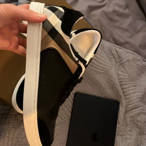 Burberry sneakers med rutigt mönster - Snygga Burberry sneakers med klassiskt rutigt mönster i beige, svart och vitt på sidorna. Ovandel i svart skinn och mocka, vita sulor och svarta snören. Perfekt för dig som vill ha en stilren men ändå unik look med lyxig känsla.
