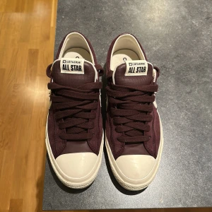 Vinröda Converse All Star sneakers - Snygga vinröda Converse All Star sneakers med klassisk vit sula och tå. Skorna har snörning, vit stjärndetalj på sidan och är tillverkade i mocka och textil. Perfekta för dig som vill ha en stilren och tidlös look med en twist. De är använda enstaka gånger och i nyskick.