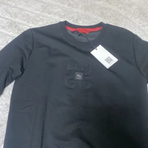 Svart sweatshirt från Hugo Boss - Svart sweatshirt  från Hugo Boss med diskret broderad HUGO-logga på bröstet. Tröjan har rund halsringning och långa ärmar. Klassisk och stilren design med röd detalj på insidan av kragen. Perfekt för dig som gillar minimalistisk stil. Storlek L men passar också till M