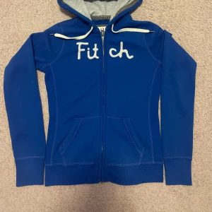 Blå hoodie från Abercrombie & Fitch - Snygg blå hoodie från Abercrombie & Fitch med vit broderad text på bröstet. Tröjan har dragkedja, huva med vit snörning och två fickor framtill. Perfekt för en chill och sportig look.