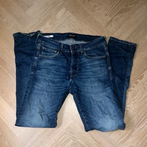 Slim Glenn jeans från Jack & Jones - Snygga blå jeans från Jack & Jones, modell Slim Glenn. De har en slim passform och klassisk femficksdesign. Jeansen är i denim med snygga slitningar och kontrastsömmar. Perfekta för dig som gillar en smal siluett och stilren look.