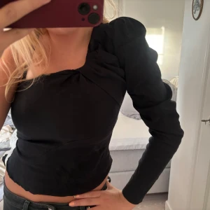 Svart oneshoulder topp med puffärm - Säljer en svart oneshoulder topp med en lång puffärm och snygga draperade detaljer vid axeln. Toppen är croppad och har en stilren, tight passform som framhäver figuren. Perfekt för dig som gillar trendiga och unika plagg med lite edge.