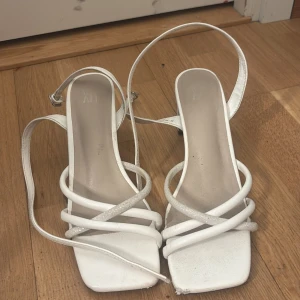 Vita sandaler med remmar från XIT - Säljer ett par vita sandaler från XIT i storlek 38. Skorna har flera tunna remmar, varav en med glittereffekt, fyrkantig tå och låg klack. De har en stilren look med öppen tå och platt sula, perfekta för sommarens alla outfits.