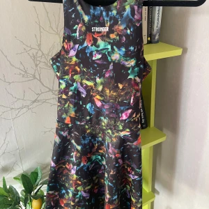 Färgglad träningsklänning från Stronger - Sportig kortklänning från Stronger med färgstarkt abstrakt mönster i svart bas och inslag av lila, grön, blå, orange och rosa. Ärmlös modell i stretchigt syntetmaterial, perfekt för dig som vill sticka ut på gymmet eller under utomhusaktiviteter. Strl S. Följer med ett par svarta shorts med ficka, som hör till klänningen. 