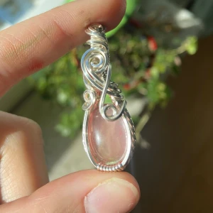 Silverberlock med rosenkvarts  - Unik berlock med en droppformad, ljusrosa sten infattad i snirklig silvertråd. Stenen har en mjuk glans och infattningen är detaljerad med vackra virvlar som ger ett bohemiskt och konstnärligt uttryck. Perfekt för dig som gillar personliga smycken med magisk känsla. Kedja 50kr extra. 