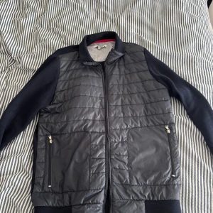 Svart quiltad jacka från Iceland M - Snygg svart quiltad jacka från Iceland i storlek M. Jackan har glansig, vadderad framsida med dragkedja och två fickor med dragkedjor. Ärmarna är i mjukare tyg och muddar vid ärmslut och midja ger en sportig look.